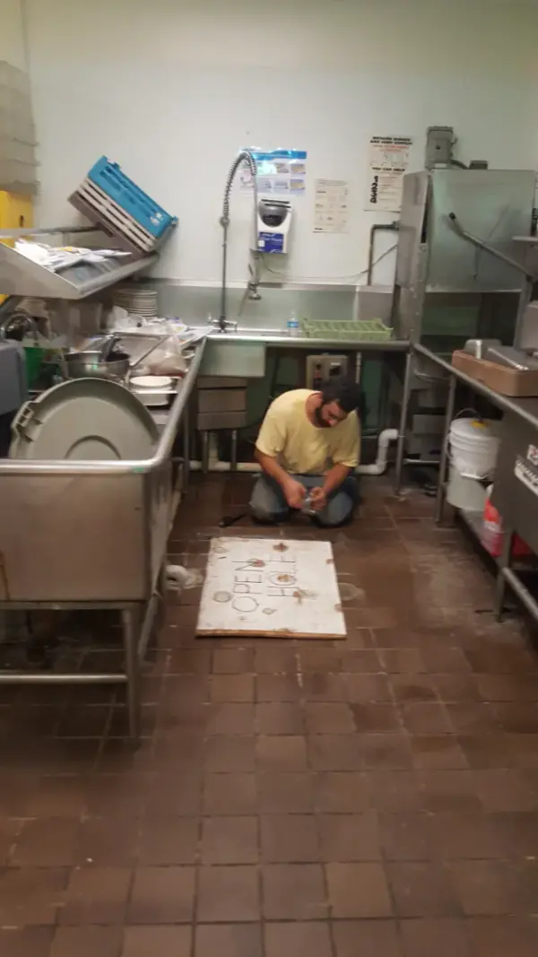 Plumber kneeling on a commercial kitchen floor beside an opened floor access panel, working on the drain line under the dish station