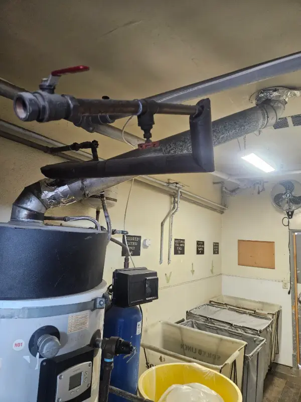 Commercial laundry room with a water heater, blue water softener tank, and overhead gas and vent piping