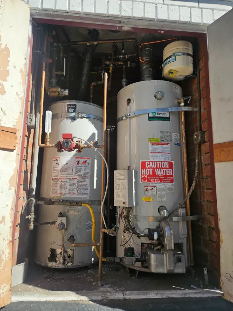 Two large commercial water heaters with straps, copper piping and an expansion tank in an outdoor closet