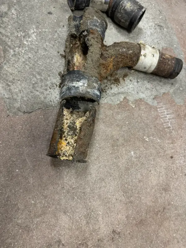 Corroded metal pipe junction with rust and taped sections on a concrete floor