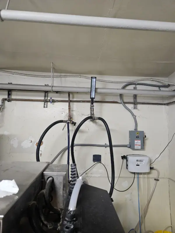 Overhead water piping, a thermometer and a Butler chemical dispenser above a commercial dishwasher