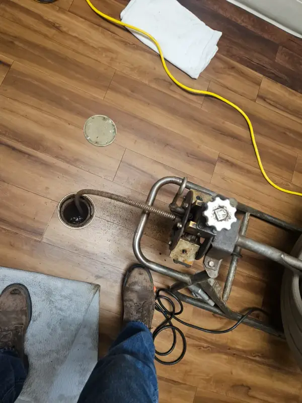 Drain machine feeding cable into a floor cleanout opened in a wood-plank floor
