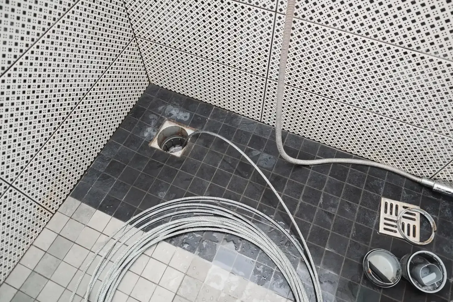Drain snake cable fed into an open shower floor drain, with the grate and drain parts set aside