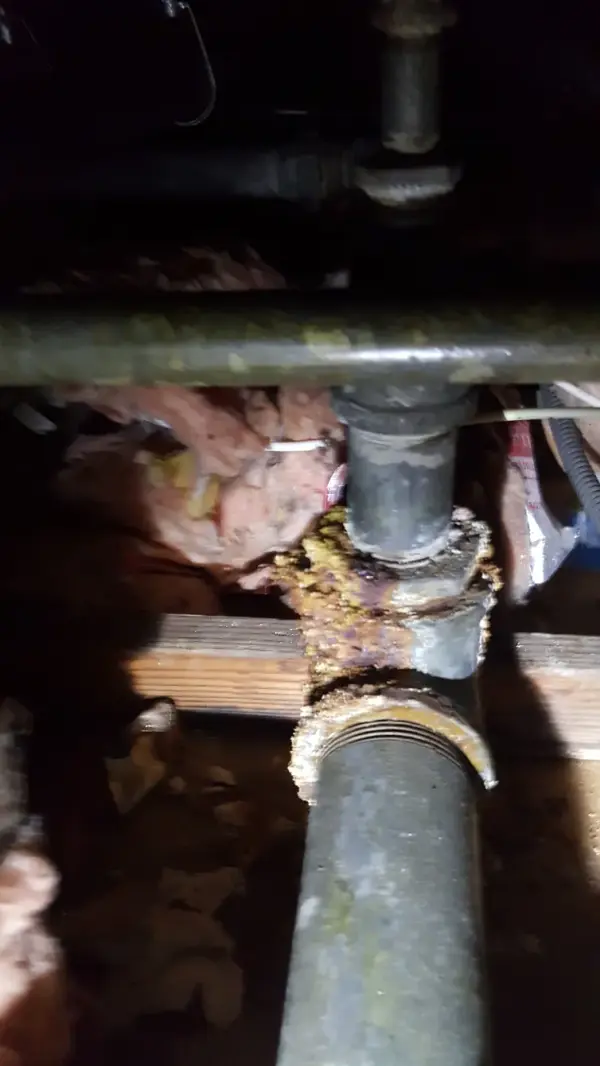 Corroded galvanized pipe joint leaking in a crawl space, with mineral buildup crusted around the fitting