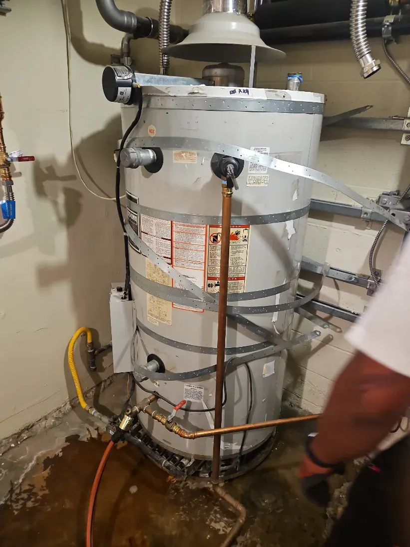 Strapped gas water heater on a wet utility room floor, with a plumber's gloved arm at the right