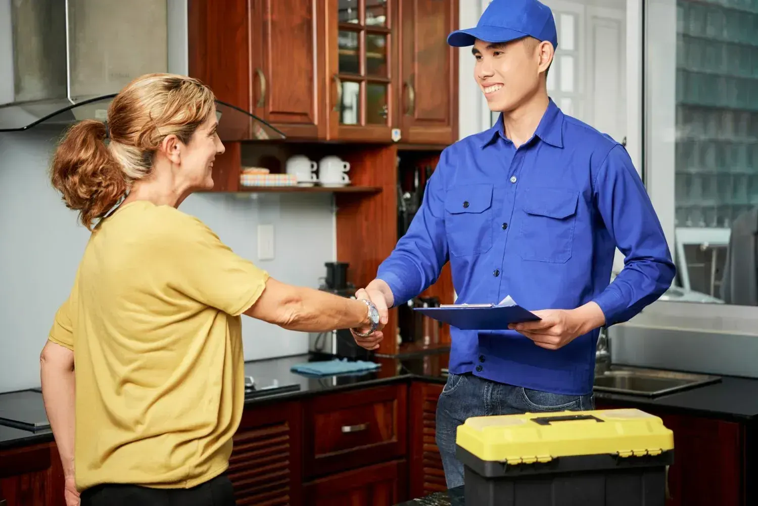 Plumber in uniform shaking hands with a homeowner in her kitchen, toolbox on the counter