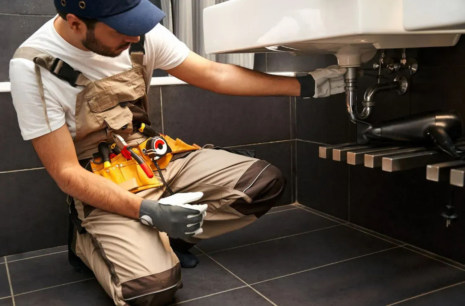 Kneeling plumber with a tool belt checking the P-trap under a wall-mounted bathroom sink