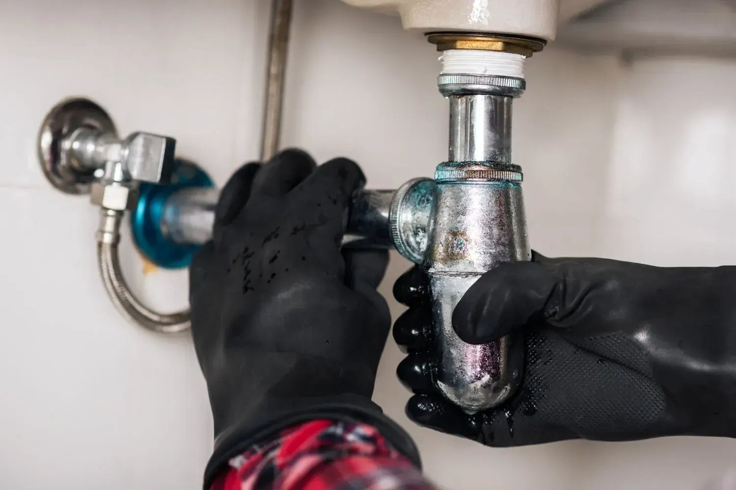 Hands in black rubber gloves loosening a chrome bottle trap under a bathroom sink