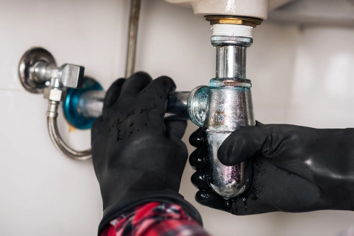 Hands in black rubber gloves removing a corroded chrome bottle trap under a sink