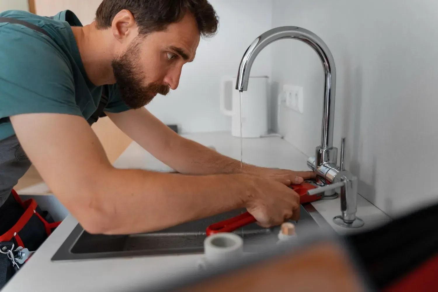 Plumber tightening a kitchen faucet with a red pipe wrench while water runs from the spout