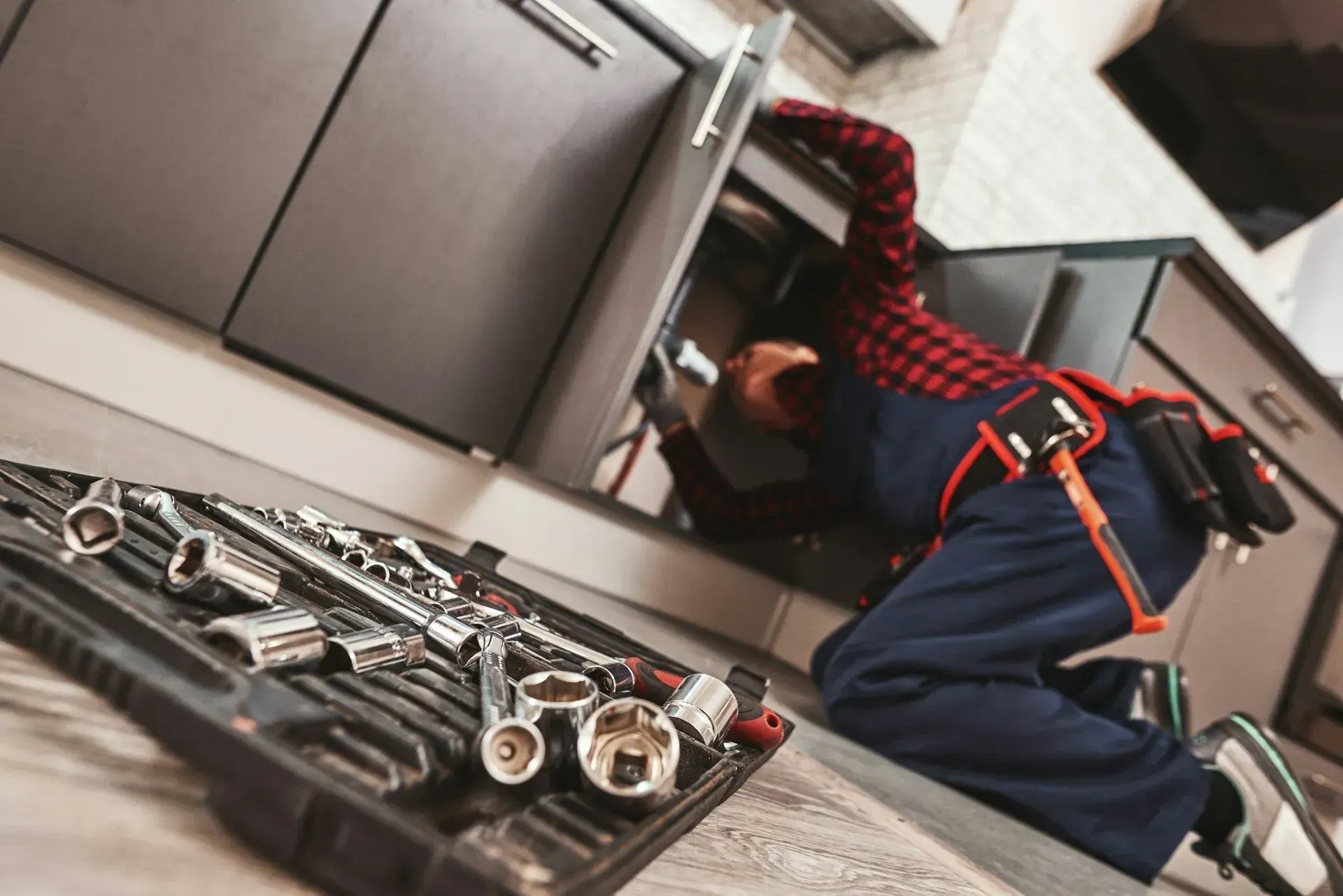 Plumber working under a kitchen sink beside an open case of socket wrenches