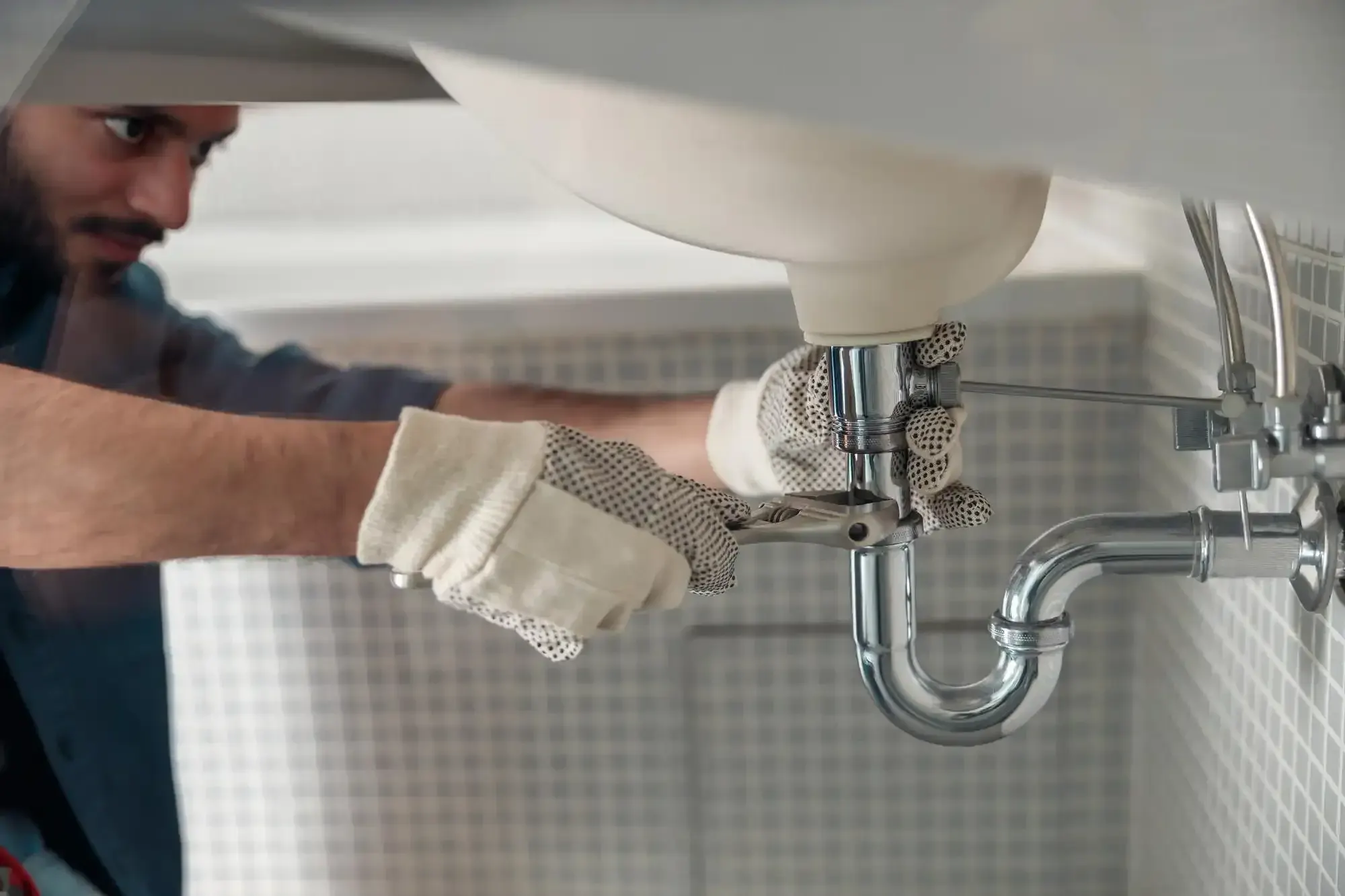 Plumber in dotted work gloves tightening a chrome P-trap under a bathroom sink with a wrench