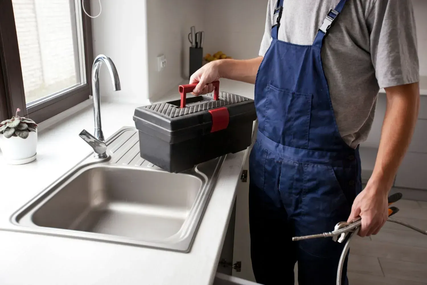 Plumber in blue overalls setting a toolbox beside a kitchen sink, holding a drain cable and wrench