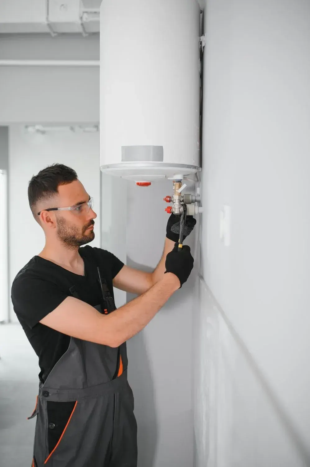 Plumber in safety glasses tightening a valve under a wall-mounted electric water heater