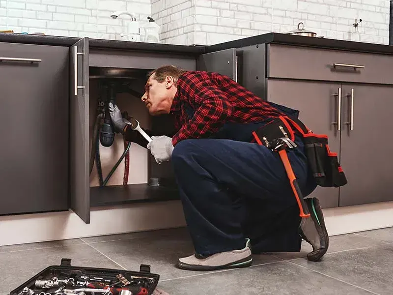 Plumber kneeling at an open kitchen cabinet, using a wrench on the pipes under the sink