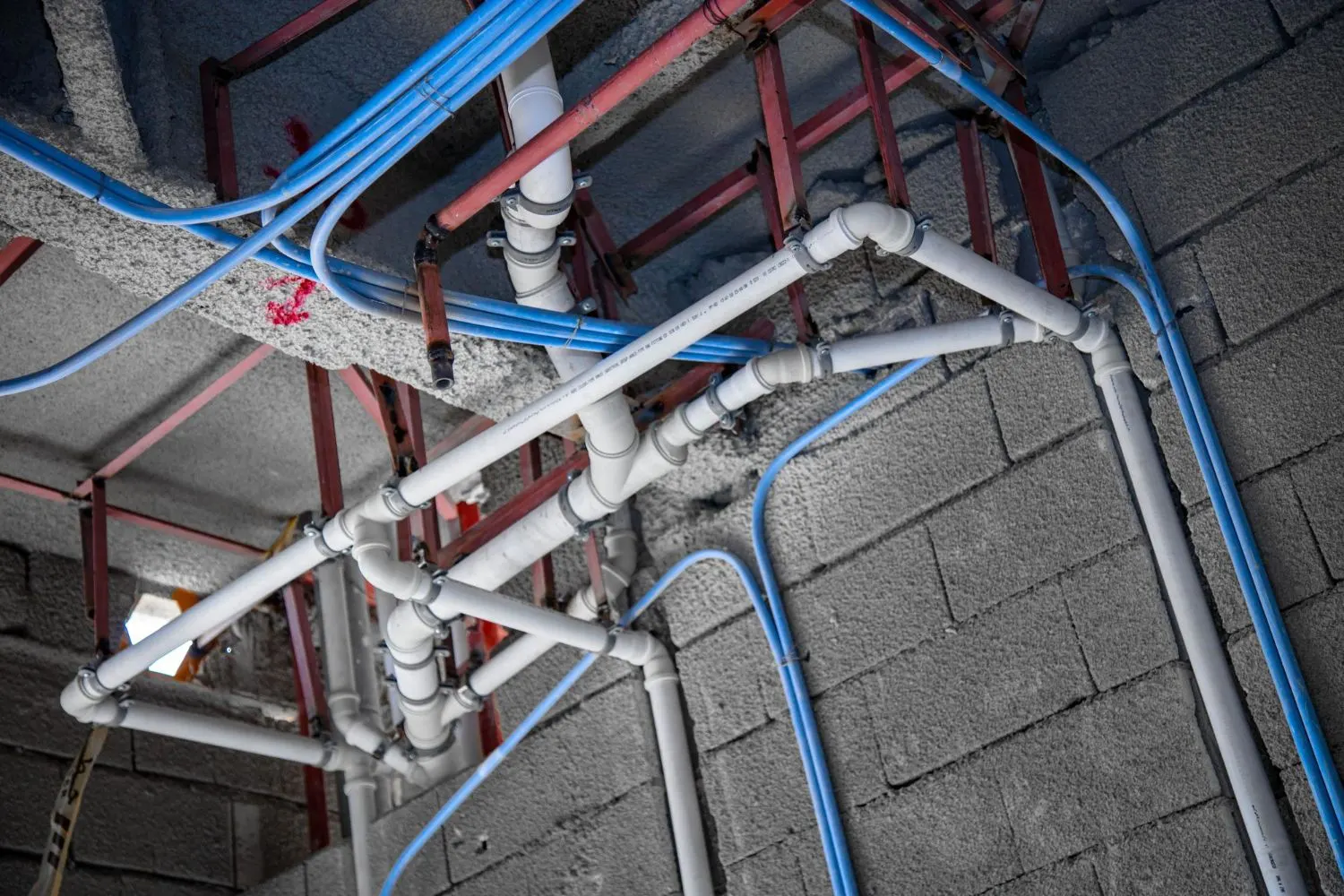 White PVC drain lines and blue water supply lines run along a block wall and ceiling during construction