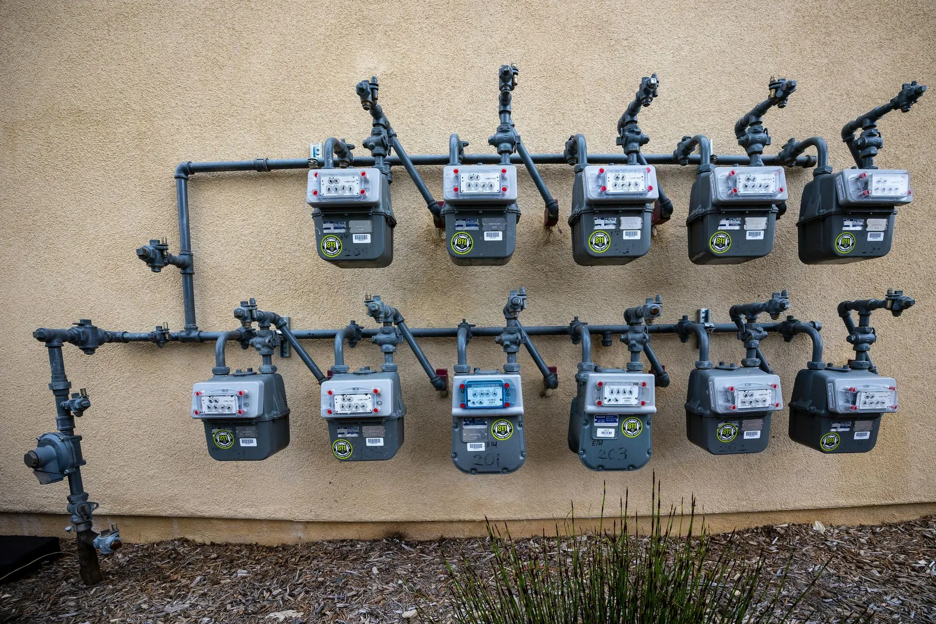 Two rows of residential gas meters on a shared manifold mounted to a stucco wall