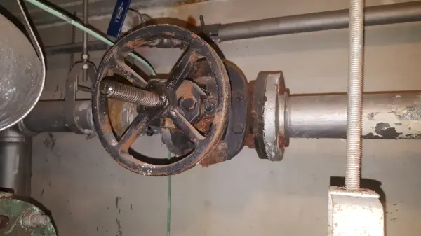 Industrial pipe with a large rusted valve wheel mounted on a metal pipeline in a utility room