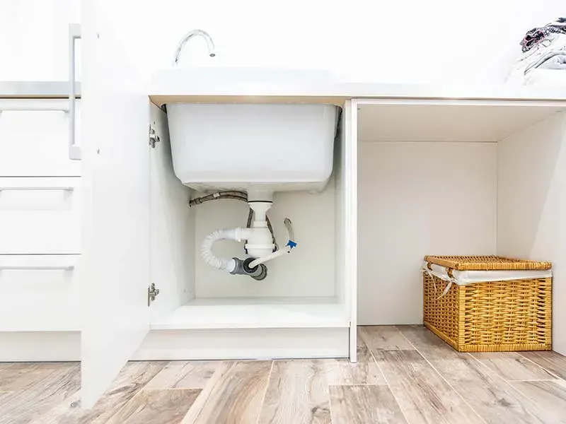 Open cabinet showing a white sink with its drain trap and flexible supply lines underneath