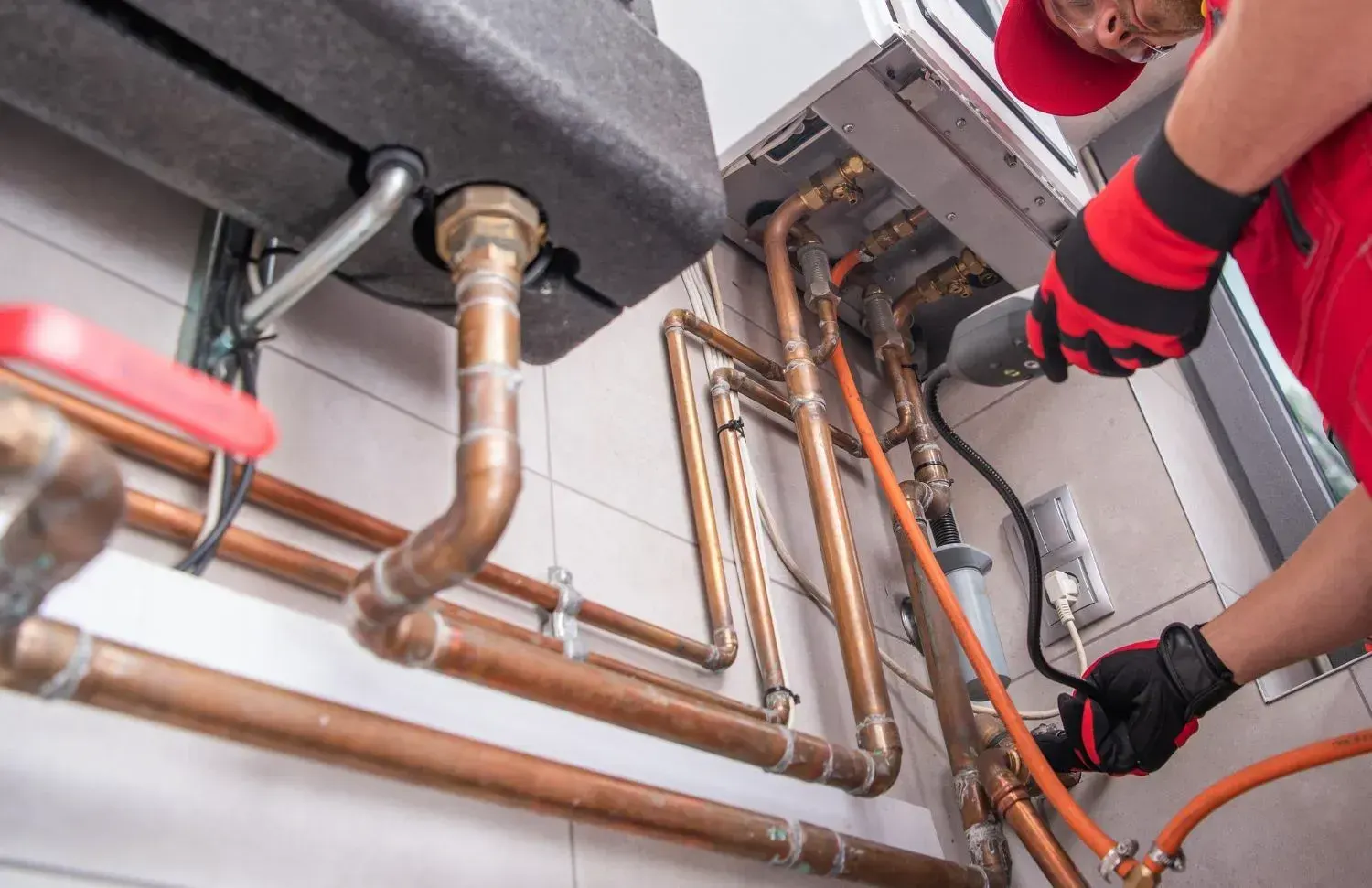 Gloved technician working on copper pipes and a gas line beneath a wall-mounted water heater