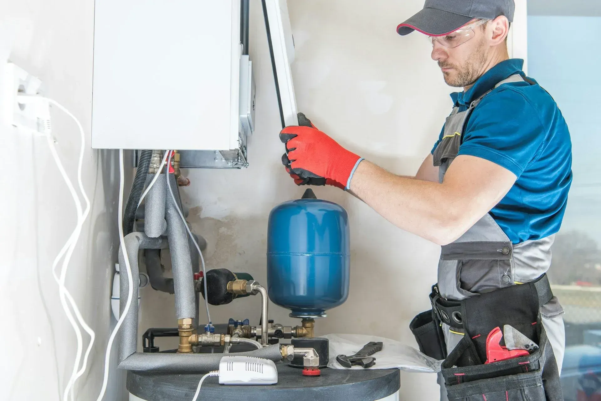 Technician in safety glasses and gloves servicing a wall-mounted boiler above a blue expansion tank