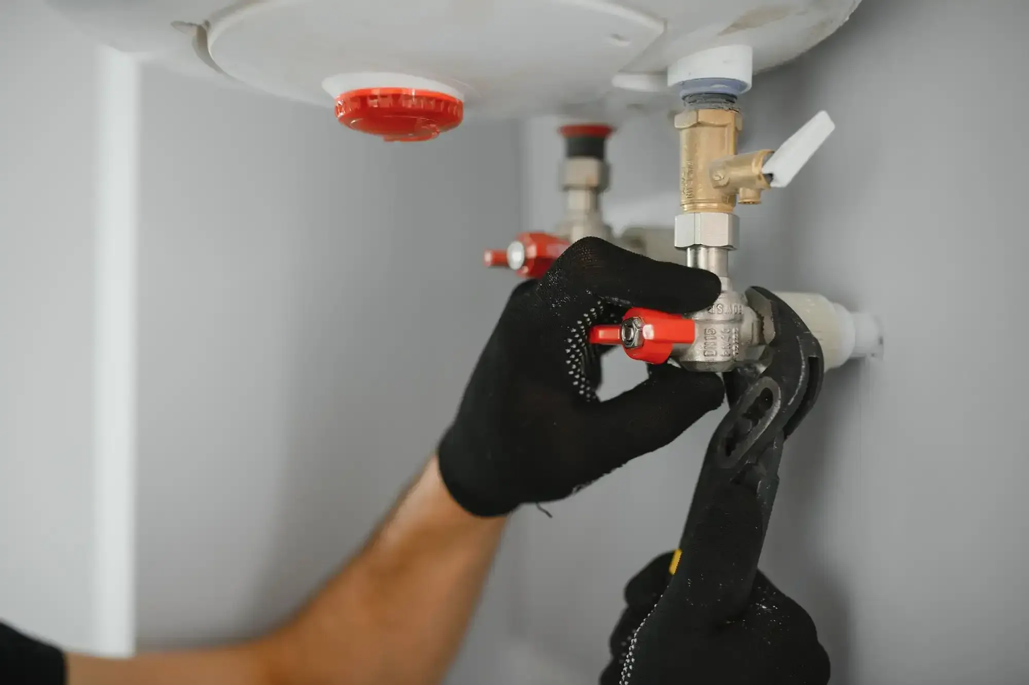 Gloved hands using pliers to tighten a shutoff valve under a wall-mounted electric water heater