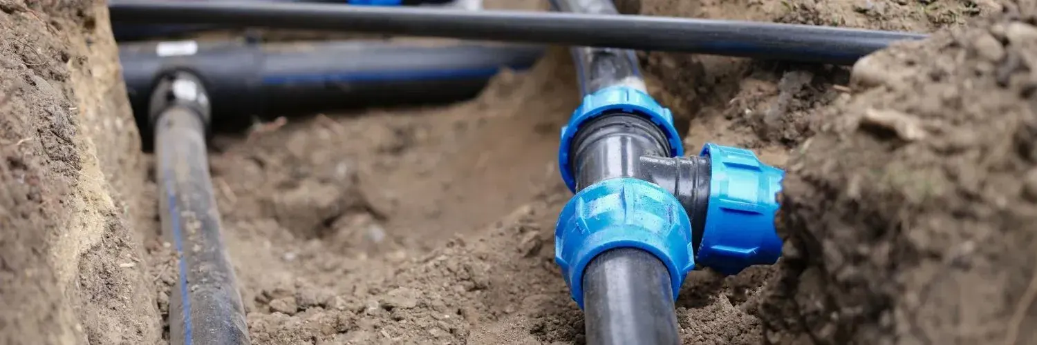 Black polyethylene water pipes joined by blue compression tee fittings in an open dirt trench